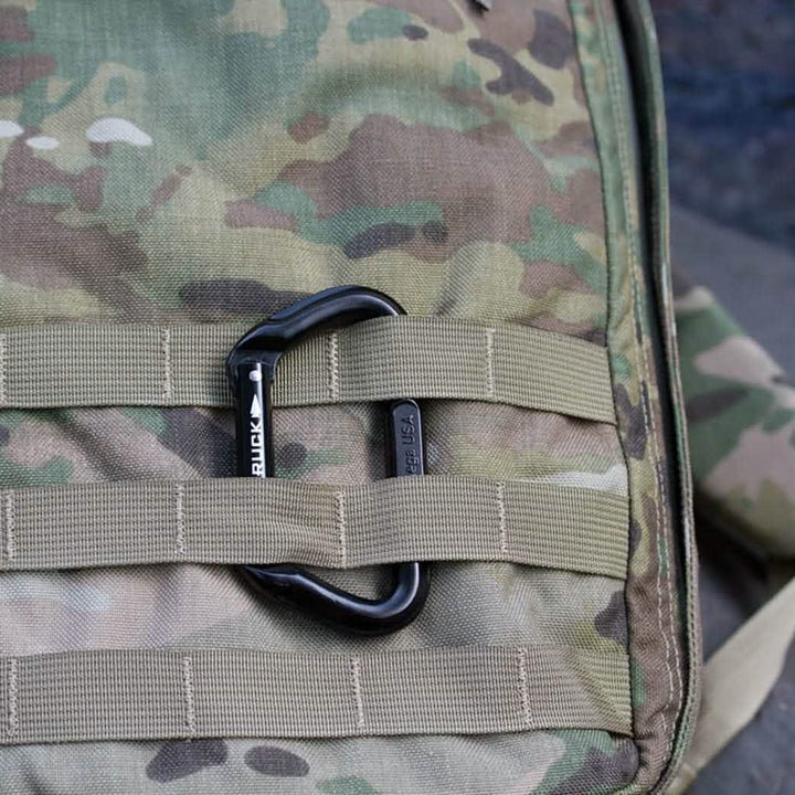 Close-up of a camouflage fabric showcasing the SMC Gear Carabiner with GORUCK Logo, attached to beige MOLLE webbing. The pattern, featuring shades of green, brown, and beige, is typically utilized in military and outdoor gear for effective concealment.