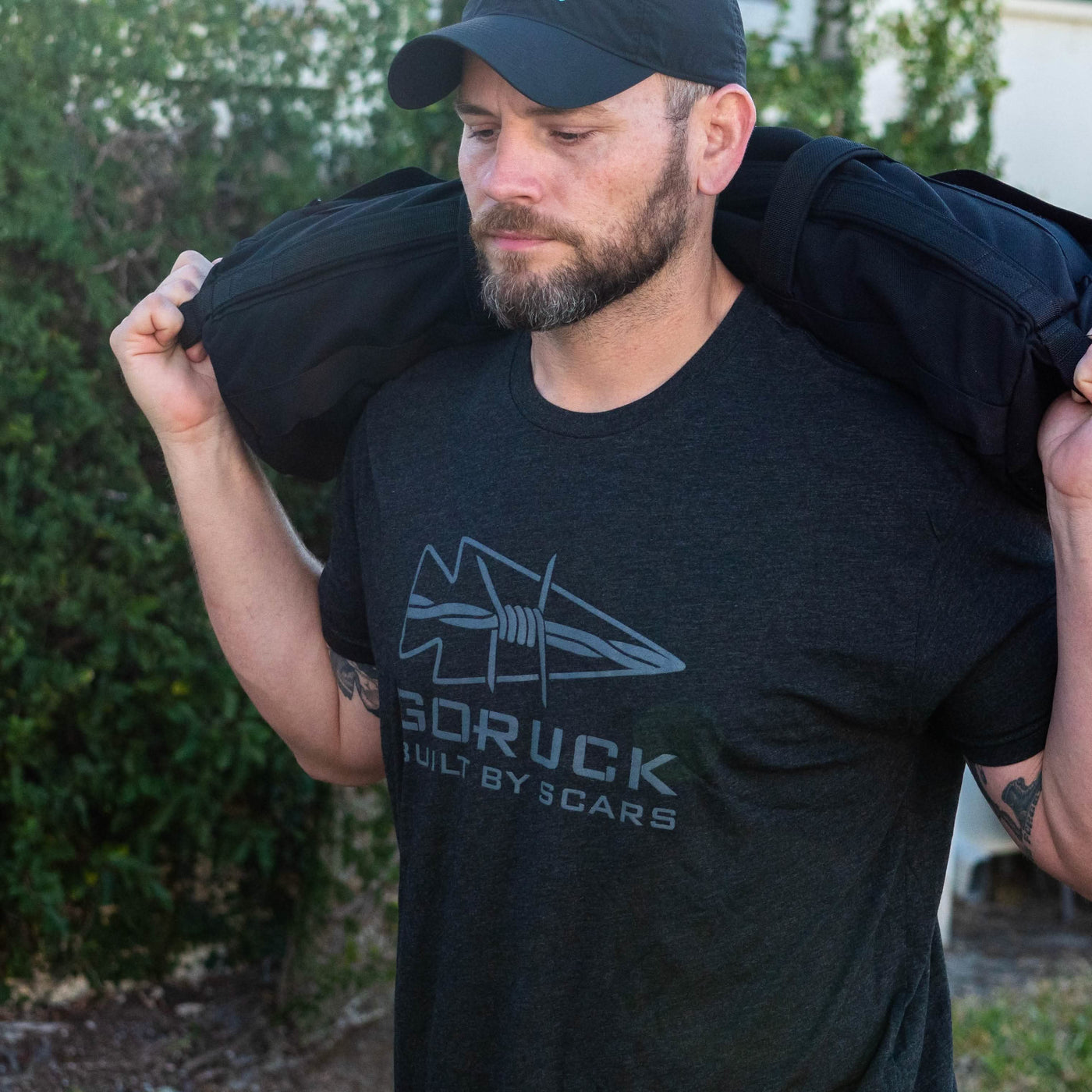 A man with a beard and a black cap carries a Frankenstein ruck on his shoulders, wearing a dark T-shirt featuring the "Built By SCARS" logo from vendor-unknown. The background showcases lush greenery.