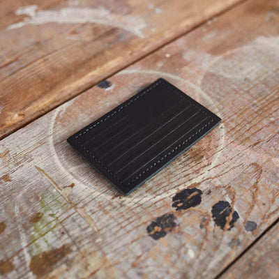 A black, genuine leather Patch - Leather Reverse Flag cardholder by vendor-unknown, featuring vertical stitching, rests on a rustic, weathered wooden surface. The paint splatters and natural wear of the wood enhance its textured appearance, complementing the cardholder's sleek design.
