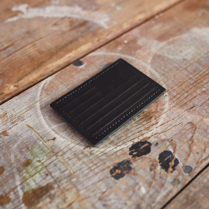 A black, genuine leather Patch - Leather Reverse Flag cardholder by vendor-unknown, featuring vertical stitching, rests on a rustic, weathered wooden surface. The paint splatters and natural wear of the wood enhance its textured appearance, complementing the cardholder's sleek design.