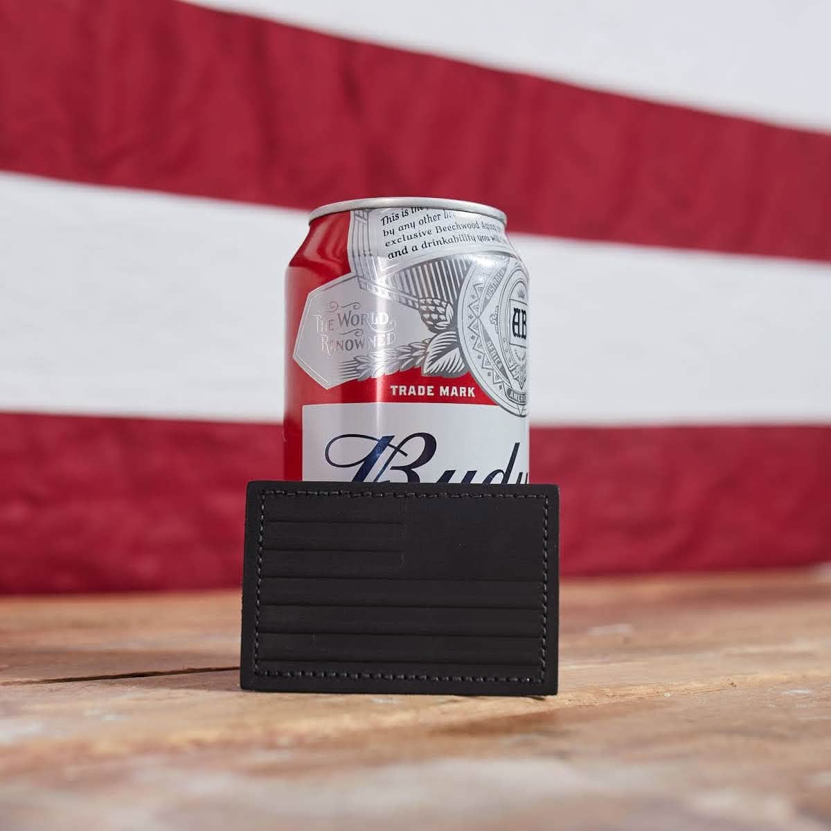 A can of Budweiser sits on a wooden table, partially covered by the Patch - Leather Reverse Flag from vendor-unknown. The red and white striped fabric in the background enhances the scene, while the genuine leather cardholder adds a touch of sophistication to this classic setup.