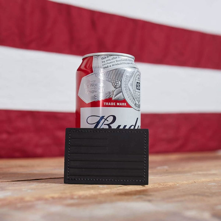 A can of Budweiser sits on a wooden table, partially covered by the Patch - Leather Reverse Flag from vendor-unknown. The red and white striped fabric in the background enhances the scene, while the genuine leather cardholder adds a touch of sophistication to this classic setup.