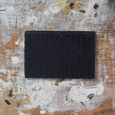 A black Patch - Leather Reverse Flag crafted with genuine leather from vendor-unknown rests on a textured wooden surface, adorned with paint splatters and scratches.