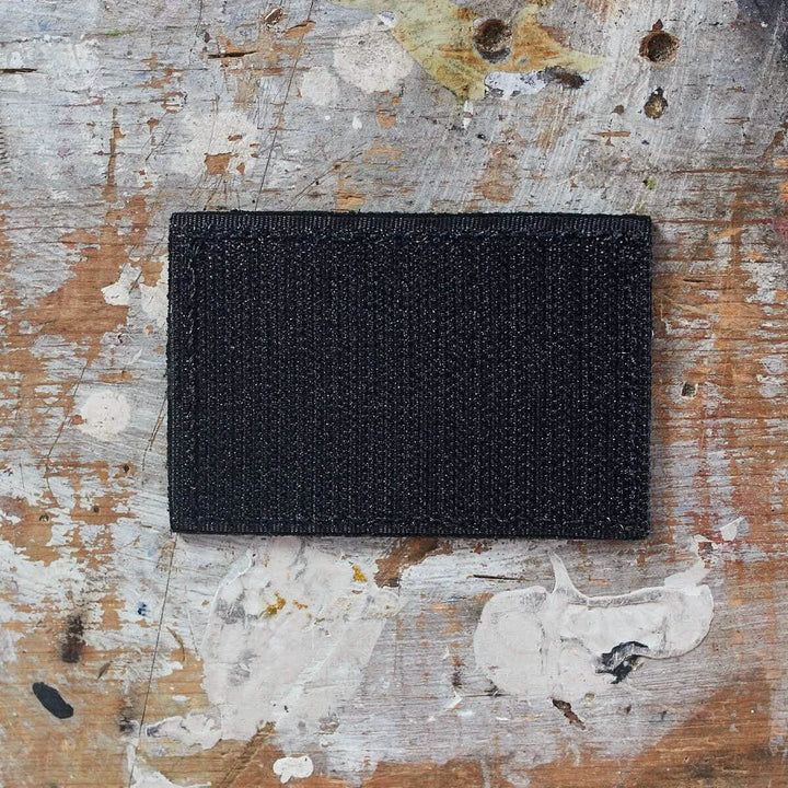 A black Patch - Leather Reverse Flag crafted with genuine leather from vendor-unknown rests on a textured wooden surface, adorned with paint splatters and scratches.