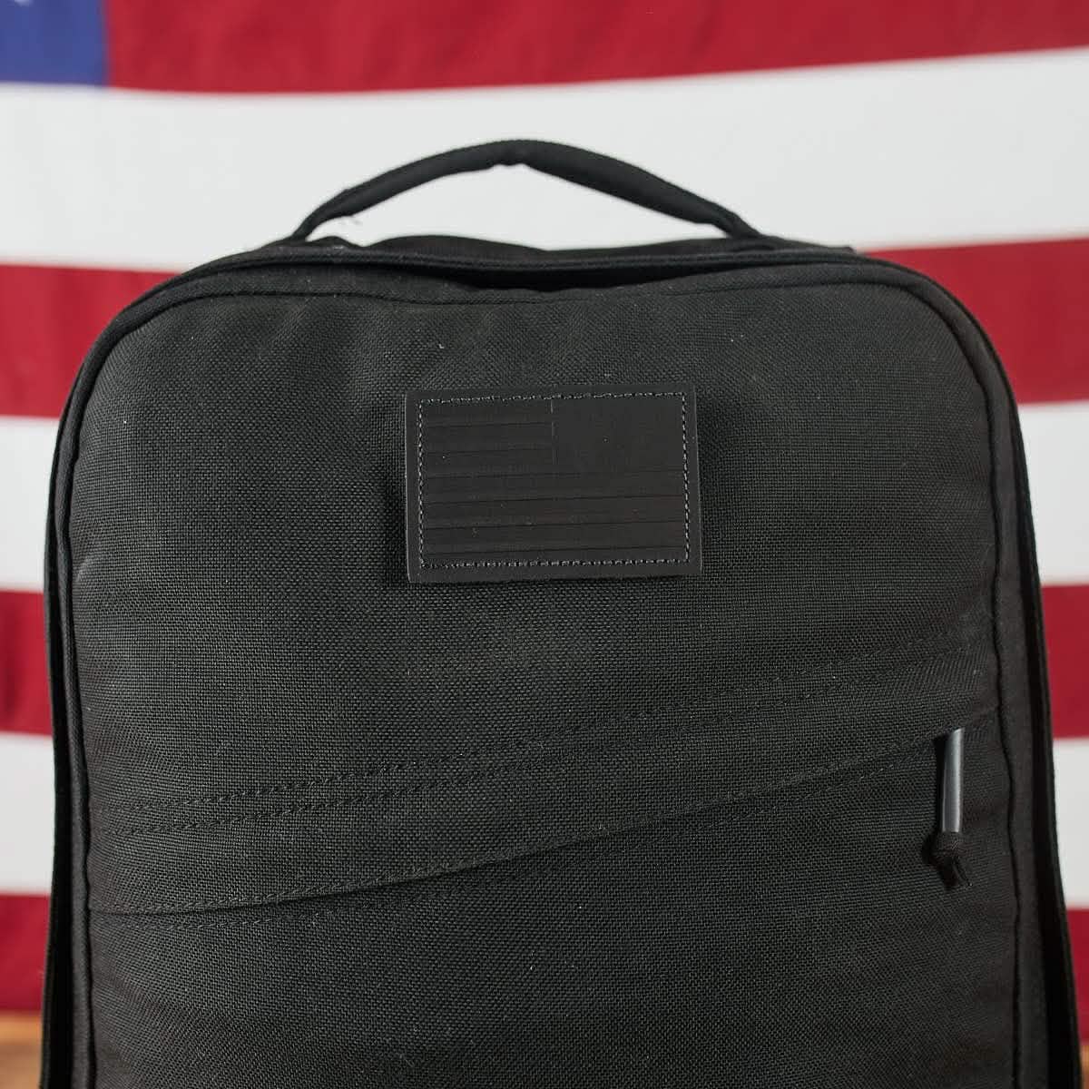 The Patch - Leather Reverse Flag backpack by vendor-unknown is a black bag with a discreet American flag patch in the center, featuring full grain cowhide VELCRO® backing. Set against a blurred American flag background, it includes a top handle and a front zippered pocket.