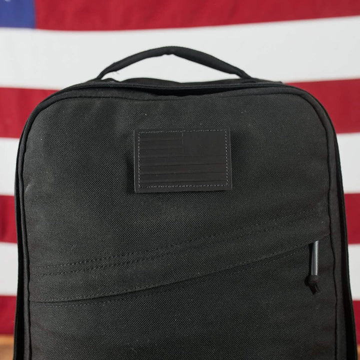 The Patch - Leather Reverse Flag backpack by vendor-unknown is a black bag with a discreet American flag patch in the center, featuring full grain cowhide VELCRO® backing. Set against a blurred American flag background, it includes a top handle and a front zippered pocket.