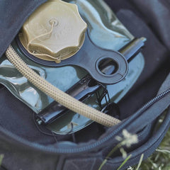 Nestled inside an open rucksack, a Nite Ize hydration bladder with a gold-colored cap and hose peeks out, suggesting the start of an exciting outdoor adventure. The surrounding grass fades out of focus, while the included Nite Ize Carabiner – Hydration Bladder Attachment hangs nearby, preparing for any rugged journey ahead.