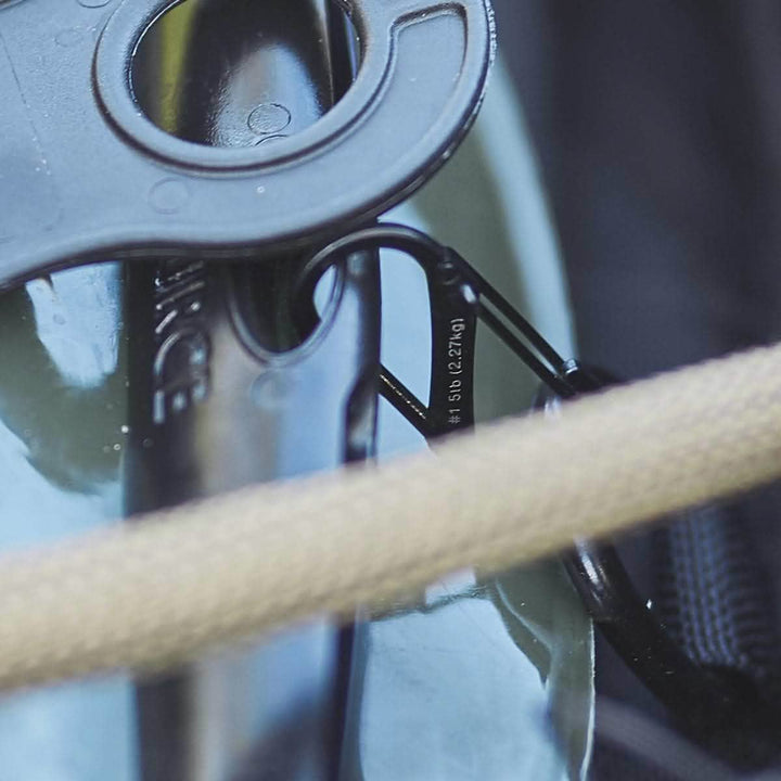 Close-up of a Nite Ize Carabiner - Hydration Bladder Attachment linked to a loop on the green surface of a rucksack. A beige strap traverses the image, indicating that the object is securely fastened.