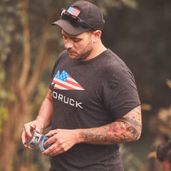 Man outdoors wearing a dark GORUCK t-shirt with American flag logo and black cap with flag patch, holding playing cards