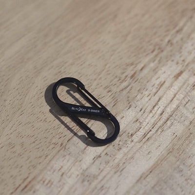 A compact black metal S-shaped carabiner sits on a wooden surface, displaying the Nite Ize brand. Ideal for attaching to your rucksack, this Carabiner - Hydration Bladder Attachment features a spring-loaded gate on each end, designed to connect items securely.