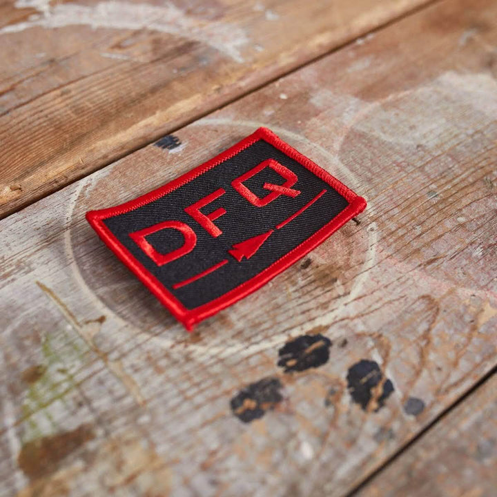 A rectangular patch from vendor-unknown, named "Patch - DFQ - Black + Red," features the letters DFQ and an arrow design with a red border. It includes a VELCRO backing for easy attachment, resting on a rustic wooden surface that highlights its motivational message against the visible grain and paint marks.