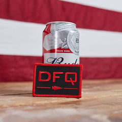 A can of Budweiser beer sits on a wooden surface, with a blurred American flag in the background. In front of the can is a vendor-unknown Patch - DFQ - Black + Red featuring an embroidered arrow pointing right and equipped with VELCRO backing for easy attachment.
