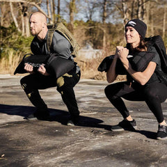Two people are outdoors doing squats on a paved surface, holding GORUCK Simple Training Sandbags and wearing casual athletic clothing. Trees and a clear sky frame their workout, making the most of their home gym in nature.