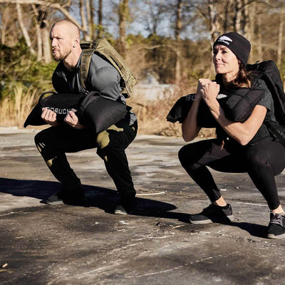 Two people are outdoors doing squats on a paved surface, holding GORUCK Simple Training Sandbags and wearing casual athletic clothing. Trees and a clear sky frame their workout, making the most of their home gym in nature.