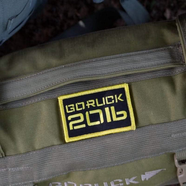 A close-up of a military-style olive green backpack adorned with a VELCRO patch labeled "Patch - Sandbag Weight" in yellow and black. The bag showcases zippers and straps, with a partially visible logo below the patch, embodying the rugged durability characteristic of high-quality rucksacks from vendor-unknown.