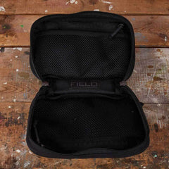 A GR1 Field Pocket by GORUCK, in an open black mesh zippered design, sits on a textured wooden surface. It reveals compartments ideal for organizing camera gear, with the text FIELD visible on the inner fabric, suggesting its rugged versatility.