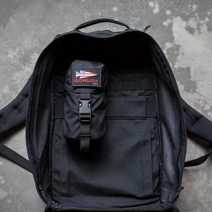 A black backpack from GORUCK with an open main compartment rests on a concrete floor, highlighting its MOLLE attachment system. Inside, a smaller black pouch is secured, displaying an American flag patch with the words "GORUCK USA." A side pocket provides convenient access to your Full Panel Water Bottle Pocket designed for Nalgene-style bottles.