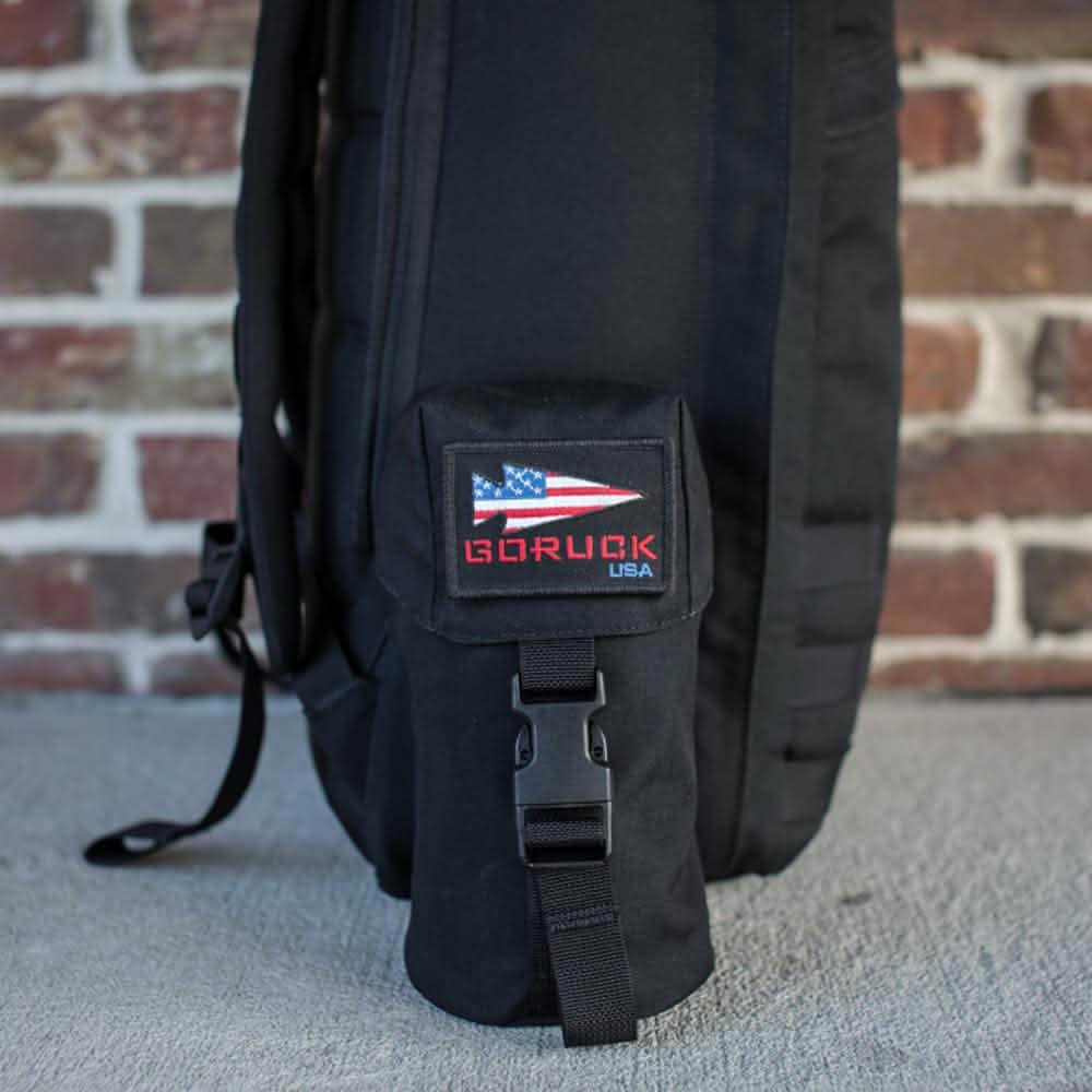 A black backpack with a Full Panel Water Bottle Pocket, adorned with a small pouch that displays an American flag patch and 
