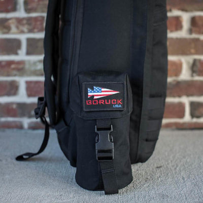 A black backpack with a Full Panel Water Bottle Pocket, adorned with a small pouch that displays an American flag patch and "GORUCK USA" in red, is leaning against a brick wall. The design features a MOLLE attachment system and includes a pocket suitable for Nalgene-style water bottles.