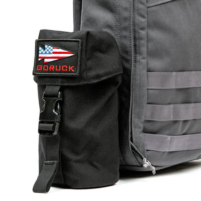 A close-up of a gray GORUCK backpack highlights its black side pouch featuring a patch with an American flag and the word GORUCK in red. With visible straps and zippers, this backpack is equipped with a MOLLE attachment system for versatile gear like the Full Panel Water Bottle Pocket designed for Nalgene-style bottles.