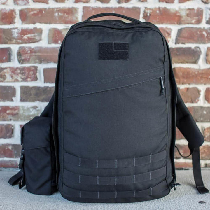A sleek black backpack with multiple compartments and a minimalist design from GORUCK is placed against a rustic brick wall. Featuring the Full Panel Water Bottle Pocket in a Nalgene style, it also boasts a matching black pouch attached to the side for extra storage.