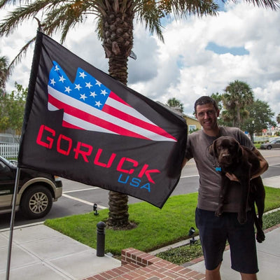 A man stands outdoors holding a "Flag - GORUCK USA," designed by Annin Flagmakers, with a large, black, stylized American theme crafted from weather-tough nylon. Next to him is a brown dog, and he smiles at the camera. Palm trees and cars are visible in the background.
