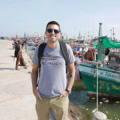A man stands smiling on a dock, wearing the GORUCK USA Tee - Tri-blend by vendor-unknown, with colorful fishing boats in the background.