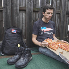 Man wearing a GORUCK t-shirt sitting on the ground eating pepperoni pizza with GORUCK backpack and boots nearby