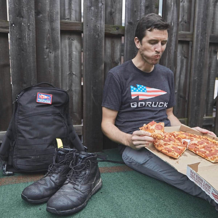 A man sits on the ground next to a wooden fence, savoring a slice of pepperoni pizza from a box. A black backpack and worn-out boots are beside him. He's wearing a dark GORUCK USA Tee - Tri-Blend by vendor-unknown and appears to be thoroughly enjoying his meal.