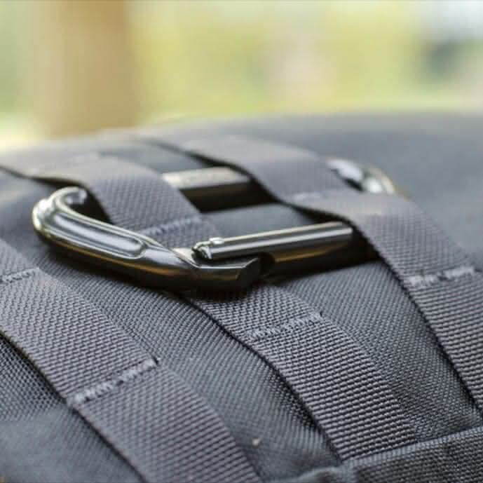 Close-up of a black aluminum carabiner clipped onto a backpack with black straps. The focus is on the GORUCK Logo carabiner by SMC Gear, showcasing the texture of the fabric and metal against a blurred background.