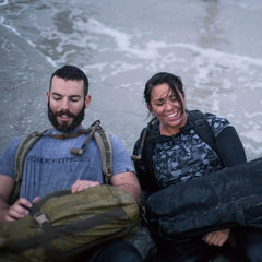 Man and woman with rucksacks sitting on wet sand, smiling after GORUCK event on beach