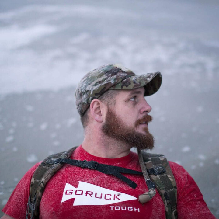 A bearded man in a camouflage cap and a backpack featuring the GORUCK Hydration Valve Mouthpiece is standing on a sandy beach. He is wearing a red GORUCK shirt with "TOUGH" printed on it, while ocean waves crash behind him.