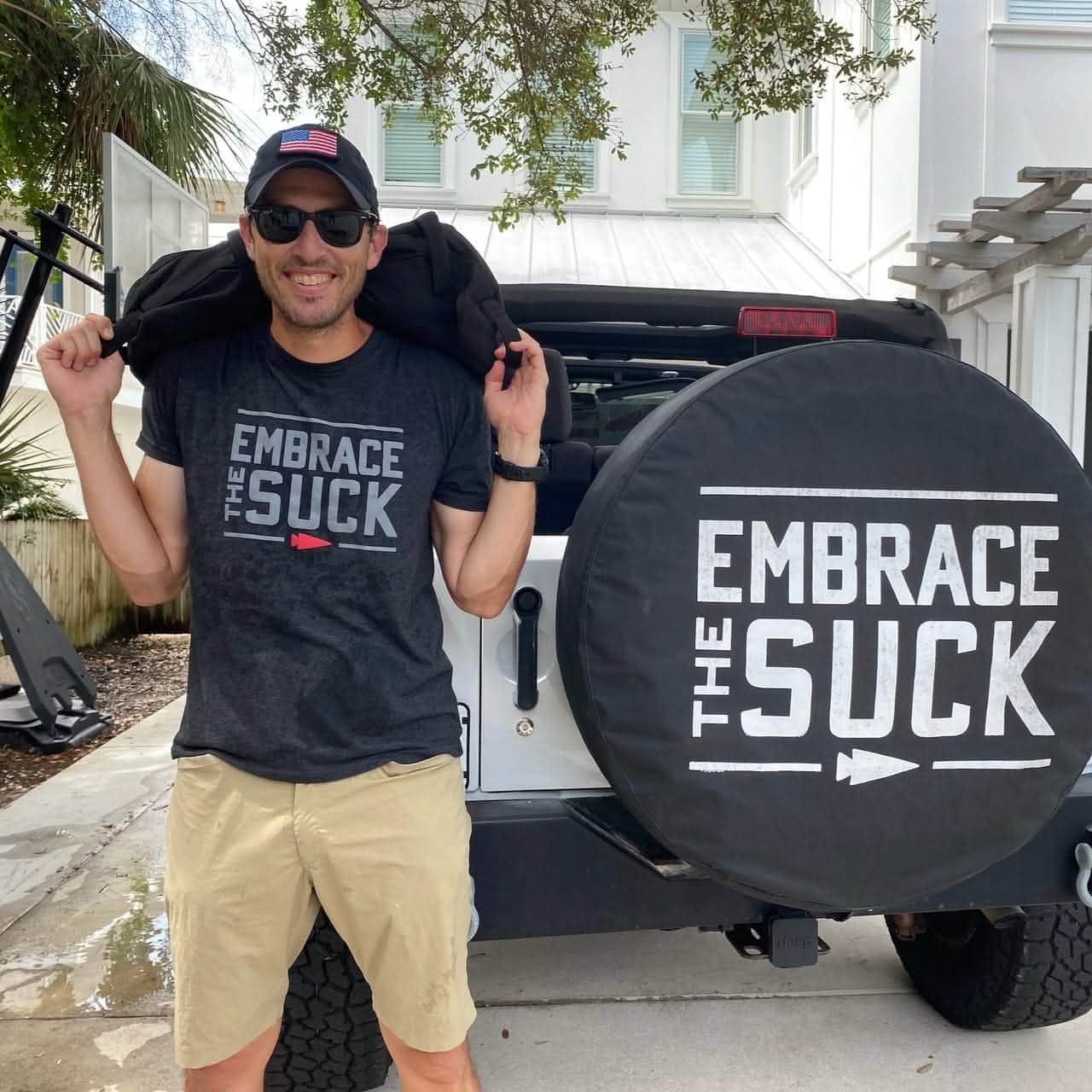 A person in a black cap and sunglasses stands next to a white Jeep, featuring a rear tire cover emblazoned with 
