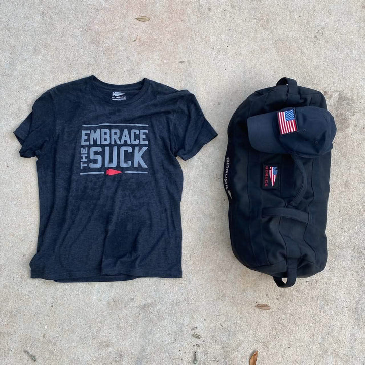 An Embrace the Suck Tee - Tri-Blend by vendor-unknown, featuring bold letters, a black cap with an American flag patch, and a black duffel bag are laid out on a concrete surface.