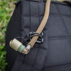 Close-up of a tan Bladder Hose Retainer from GORUCK, attached to a black strap on a backpack with a small black clip. The end features green and beige tones reminiscent of those seen at GORUCK Events. In the background, blurred green grass adds to the natural setting.