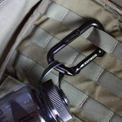 Black GORUCK branded carabiner attached to olive green ruck sack webbing with a black water bottle nearby