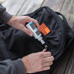 A person uses Gear Aid's Zip Care - Zipper Cleaner & Lubricant from a small bottle on the zipper of a black bag featuring an orange logo. This bag, potentially ready for GORUCK Events, is set on a wooden surface, while the person's hands are dressed in a dark long-sleeve shirt.