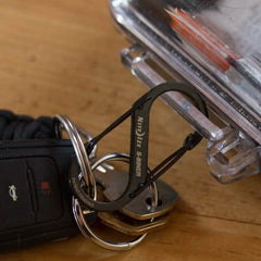 A set of keys with a black car key fob rests on a wooden table. The keys are held together by a sleek black Nite Ize Carabiner - Hydration Bladder Attachment, which is cleverly attached to a clear plastic container, much like how you might secure essentials to your rucksack for an adventure.