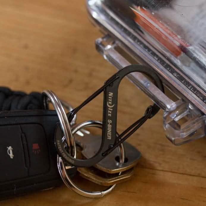 A set of keys with a black car key fob rests on a wooden table. The keys are held together by a sleek black Nite Ize Carabiner - Hydration Bladder Attachment, which is cleverly attached to a clear plastic container, much like how you might secure essentials to your rucksack for an adventure.