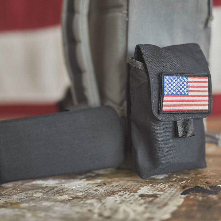 A gray backpack, designed for heavy loads, features a black pouch adorned with a U.S. flag patch and includes a GORUCK Padded Hip Belt. In front of it lies a black object, all set against the backdrop of blurred red and white stripes from an American flag.