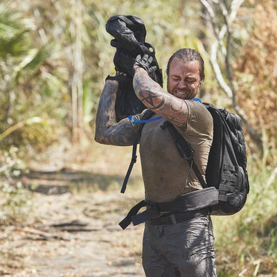 A tattooed individual is outdoors on a dirt path amidst lush greenery, dressed in a soiled shirt and pants. They are carrying a black GORUCK backpack equipped with a Padded Hip Belt, ideal for hip carry and transporting heavy loads in this rugged, natural setting.