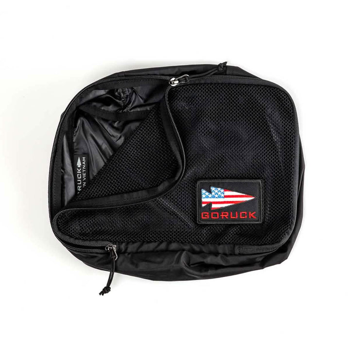 A black, partially unzipped packing cube from GORUCK, crafted from ultra-light nylon, showcases a patch with a stylized American flag and the word GORUCK. Its interior, lined with smooth and shiny fabric along with a mesh pocket, makes it an ideal travel companion for organizing essentials.