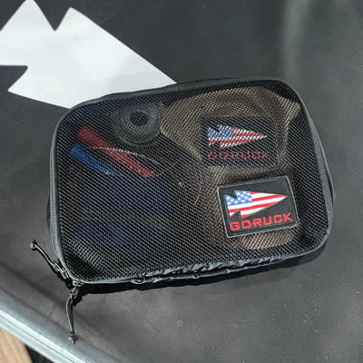 A GORUCK Packing Cube, crafted from ultra-light nylon, contains a patch featuring the American flag, a flashlight, and a folded brown item. Placed on a surface with a white arrow design, this travel companion keeps your essentials within easy reach.