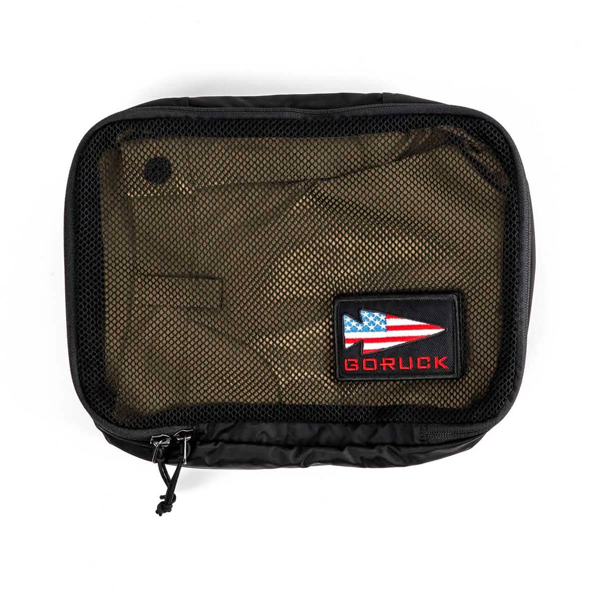 The GORUCK Packing Cubes are black travel pouches crafted from ultra-light nylon, featuring a transparent mesh front and adorned with an embroidered patch. The patch showcases a stylized red, white, and blue arrow design alongside the word "GORUCK" in red. This versatile travel companion is equipped with a zipper that includes two pull tabs.