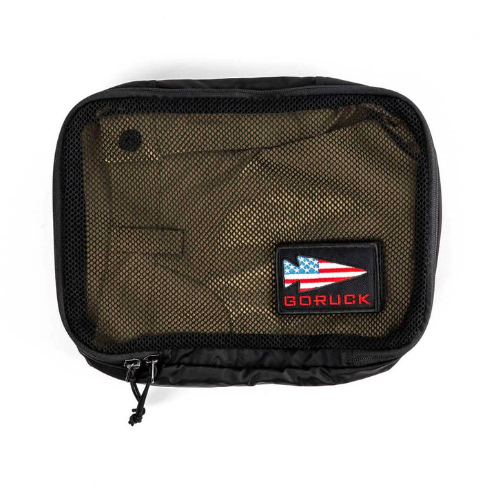 The GORUCK Packing Cubes are black travel pouches crafted from ultra-light nylon, featuring a transparent mesh front and adorned with an embroidered patch. The patch showcases a stylized red, white, and blue arrow design alongside the word "GORUCK" in red. This versatile travel companion is equipped with a zipper that includes two pull tabs.