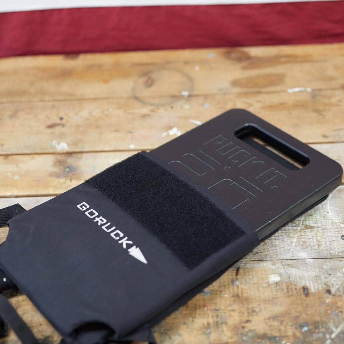 A GORUCK Ruck Plate, showcasing the excellence of American craftsmanship, is partially placed into a protective sleeve on a weathered wooden surface. In the softly focused background, an American flag subtly enhances the scene with a patriotic flair.