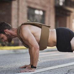 A person is performing a push-up outdoors on a paved road, wearing GORUCK's Men’s Ranger Panties - ToughStretch and a weighted vest. These shorts, crafted from ToughStretch fabric with Scars Lifetime Guarantee, provide unmatched flexibility. In the background, a brick building is illuminated by the soft glow of a sunset.