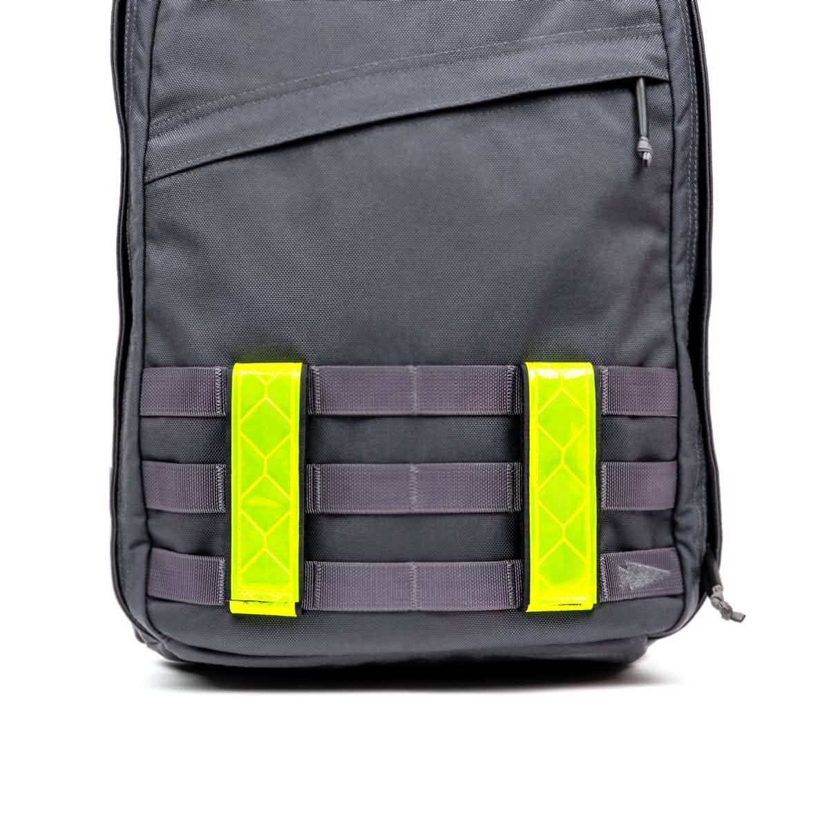 A black backpack with a minimalist design features two bright neon yellow Reflective Ruck Bands by GORUCK on the front, enhancing nighttime visibility. The backpack includes multiple horizontal loops and MOLLE webbing for attaching additional gear.