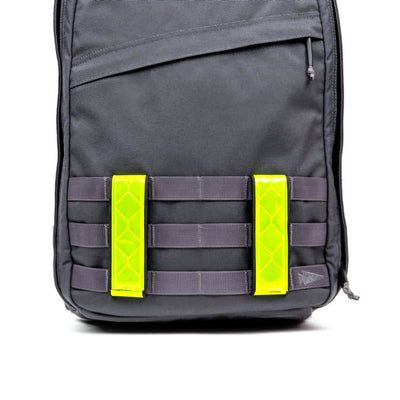 A black backpack with a minimalist design features two bright neon yellow Reflective Ruck Bands by GORUCK on the front, enhancing nighttime visibility. The backpack includes multiple horizontal loops and MOLLE webbing for attaching additional gear.