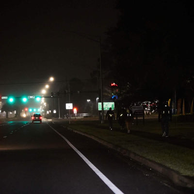 A nighttime scene shows a dimly lit road with a car driving toward a traffic light. Three people, wearing dark clothing and GORUCK Reflective Ruck Bands for nighttime visibility, walk in single file on the sidewalk. The street is illuminated by overhead lights, and the sky is overcast.